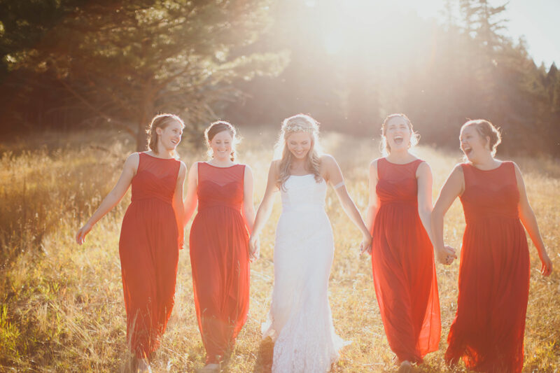 A bride walks with her bridesmaids holding hands at sunset wearing a white dress and red dresses for this Lower Lake Ranch wedding photography session