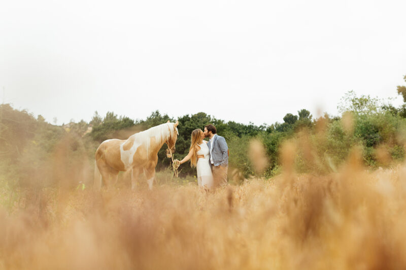 An engaged couple kiss holding each other with their horse on the hillside for this Granada Hills engagement photography session