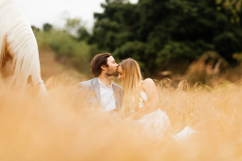 An engaged couple sit holding each other with their horse grazing in a field for this Granada Hills engagement photography session