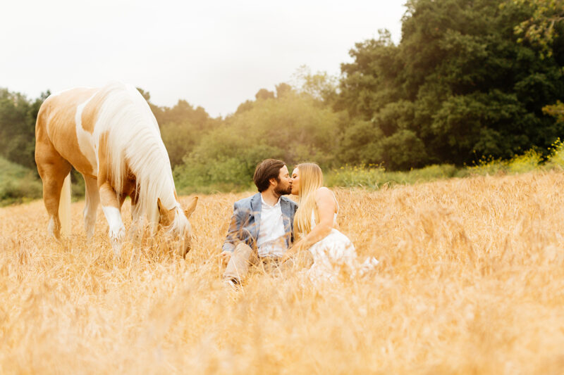 An engaged couple sit and kiss while holding each other with their horse grazing in a field for this Granada Hills engagement photography session