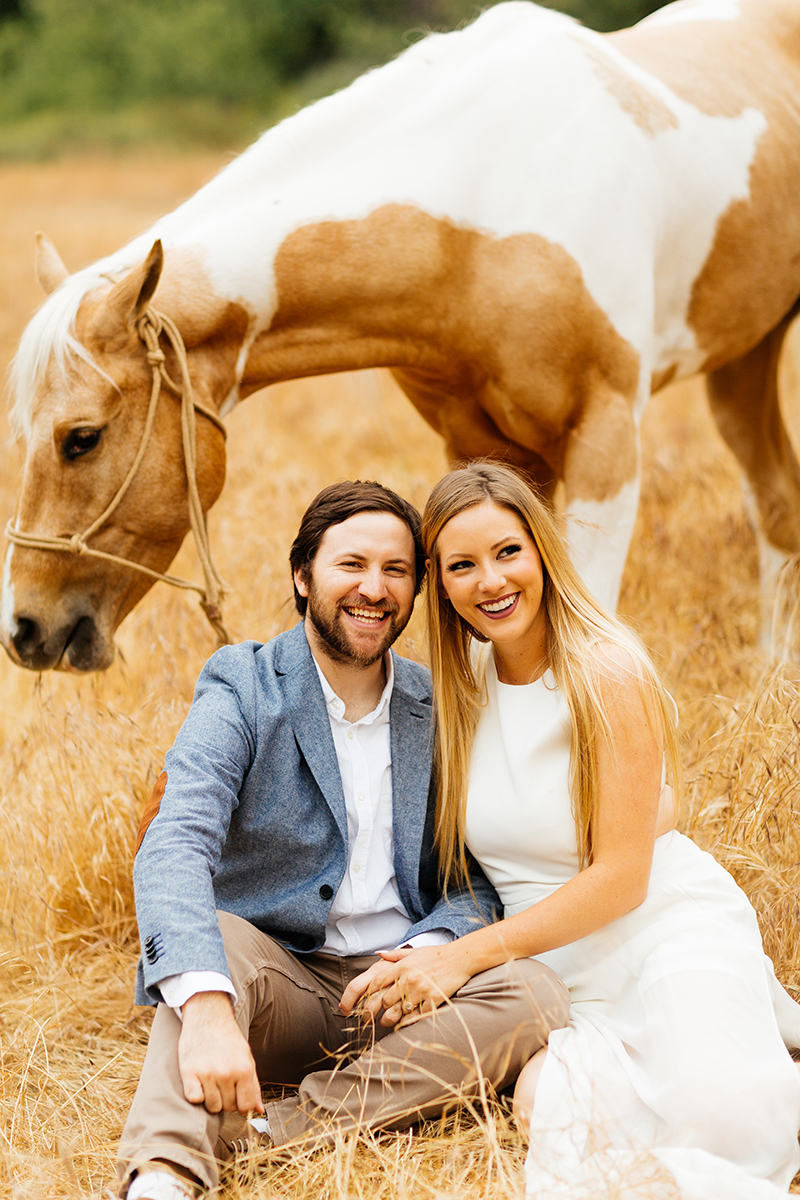 An engaged couple sit holding each other with their horse grazing in a field for this Granada Hills engagement photography session