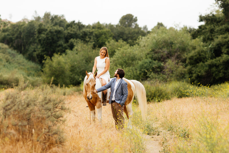 An engaged couple smile at each other as she rides their horse in a field for this Granada Hills engagement photography session