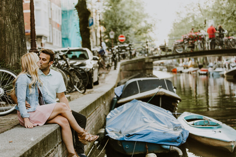 A couple sit together and kiss on the edge of a canal for this Amsterdam couples photography session