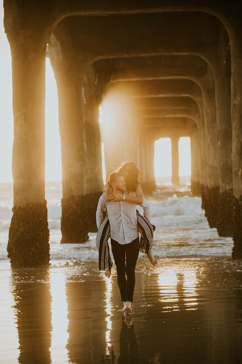 A couple walk together as he gives he a piggyback ride at sunset on the beach under a pier near the Santa Monica Pier for this Los Angeles engagement photography session