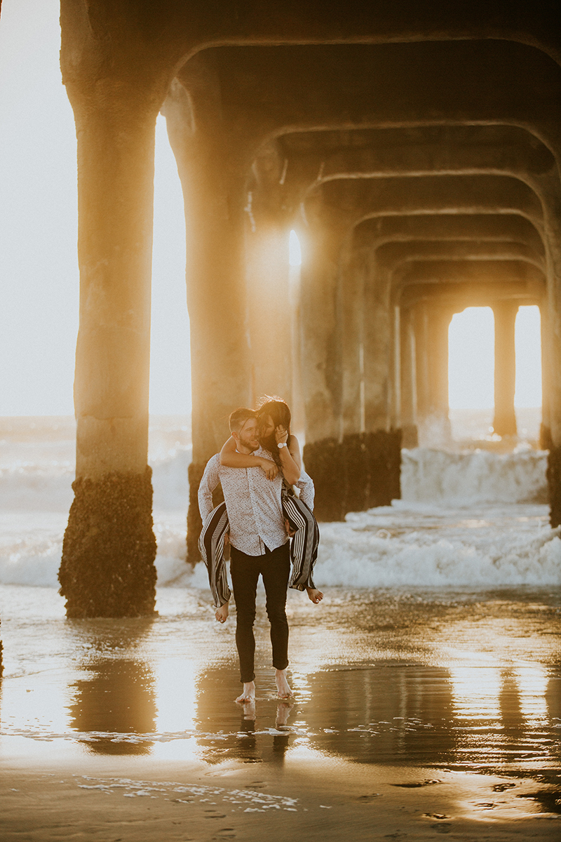 A couple walk together as he gives he a piggyback ride at sunset on the beach under a pier near the Santa Monica Pier for this Los Angeles engagement photography session