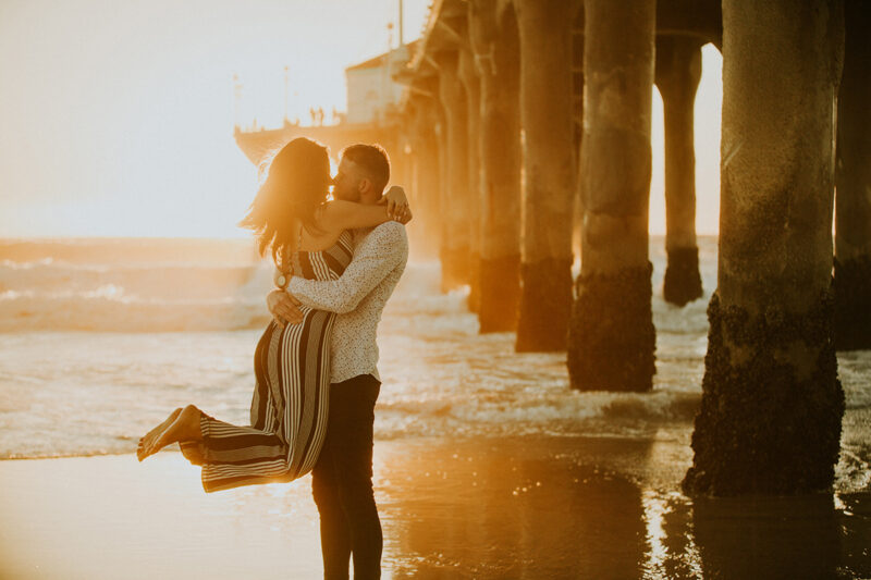A couple pose together at sunset as he lifts her up on the beach near the Santa Monica Pier for this Los Angeles engagement photography session