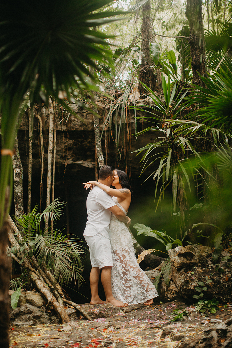 A couple hold one another kissing in front of a cenote in Mexico wearing a beautiful white dress and white shorts with a shirt for a Cenote Azul engagement photography session