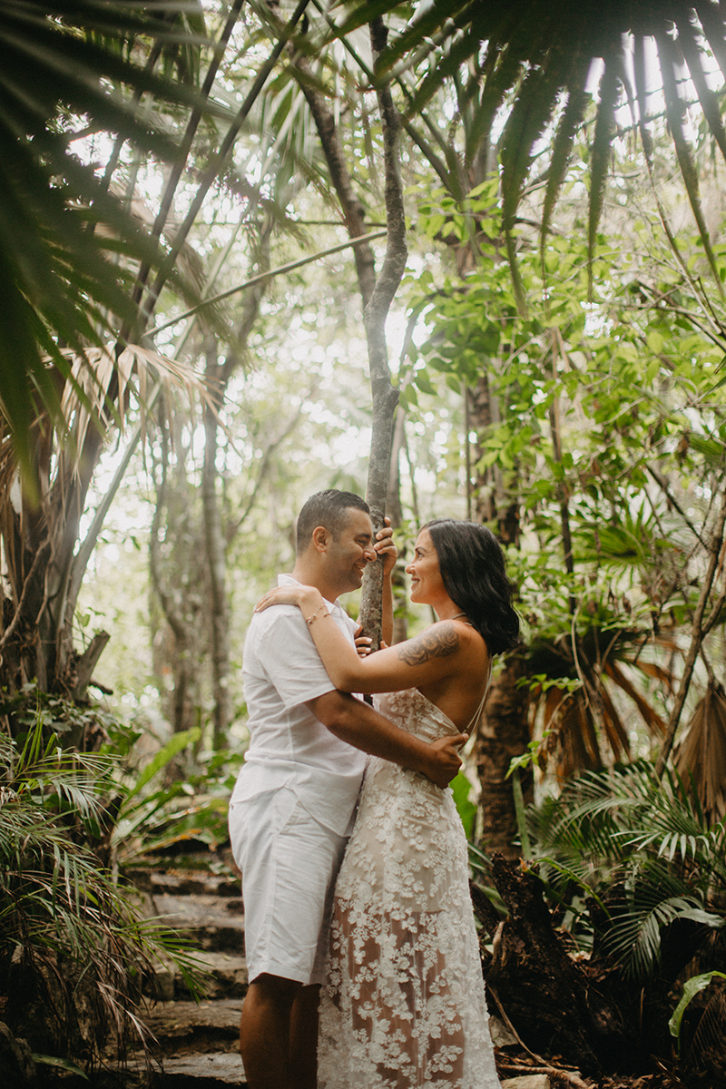 A couple hold one another smiling in front of a cenote in Mexico wearing a beautiful white dress and white shorts with a shirt for a Cenote Azul engagement photography session