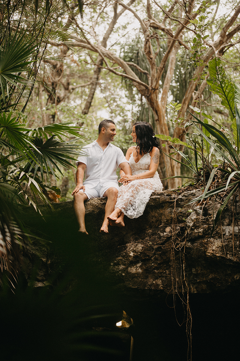 A couple sit next to each other on top of a cenote in Mexico wearing a beautiful white dress and white shorts with a shirt for a Cenote Azul engagement photography session