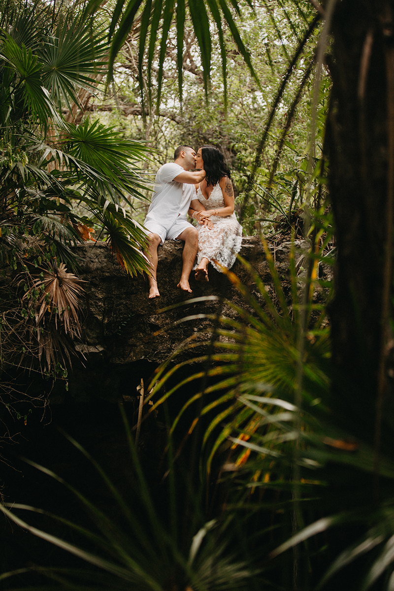 A couple sit next to each other kissing on top of a cenote in Mexico wearing a beautiful white dress and white shorts with a shirt for a Cenote Azul engagement photography session