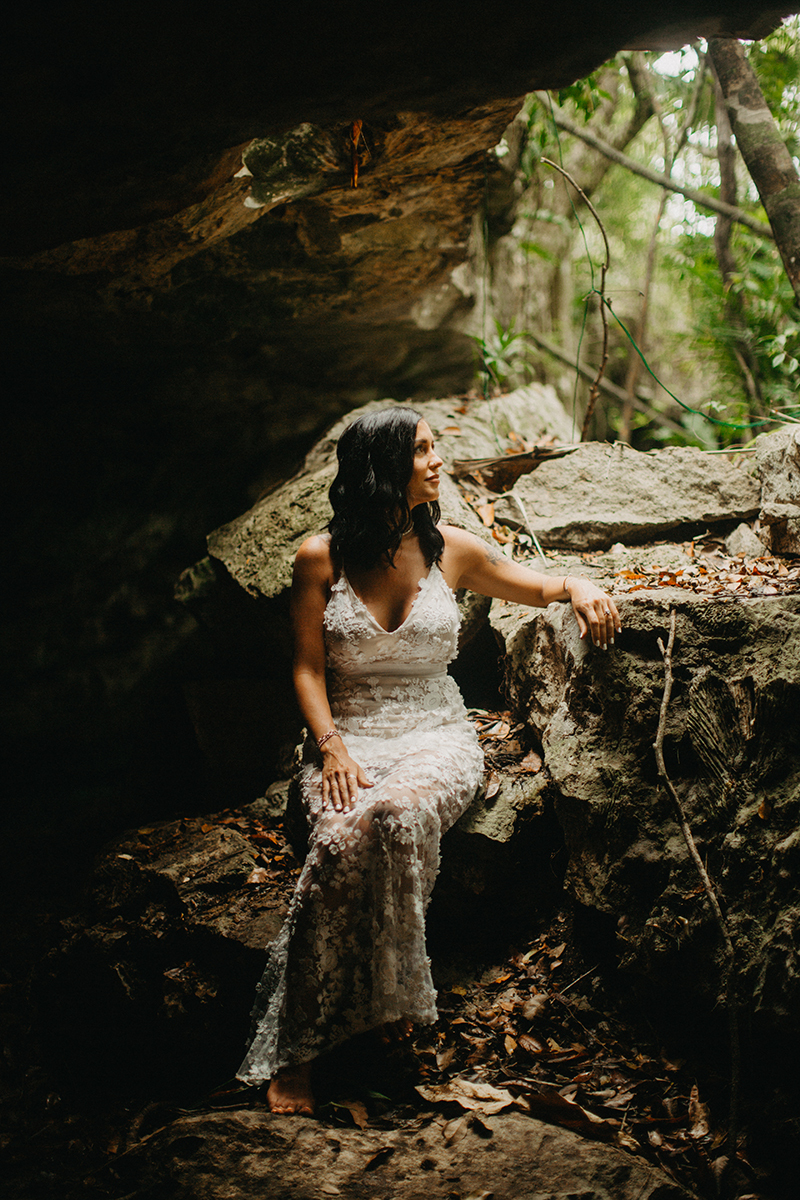 A woman poses sitting inside of a cenote in Mexico wearing a beautiful white dress and white shorts with a shirt for a Cenote Azul engagement photography session