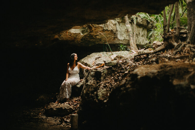 A woman poses sitting inside of a cenote in Mexico wearing a beautiful white dress and white shorts with a shirt for a Cenote Azul engagement photography session