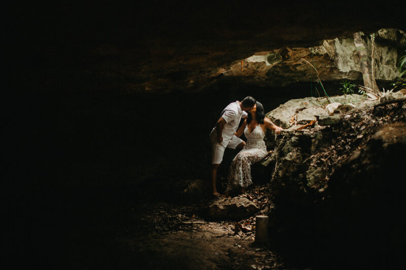 A couple together kissing inside of a cenote in Mexico wearing a beautiful white dress and white shorts with a shirt for a Cenote Azul engagement photography session