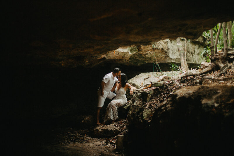 A couple together kissing inside of a cenote in Mexico wearing a beautiful white dress and white shorts with a shirt for a Cenote Azul engagement photography session