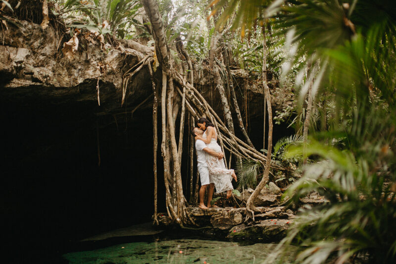 A couple stand together kissing at the edge of a cenote in Mexico wearing a beautiful white dress and white shorts with a shirt for a Cenote Azul engagement photography session