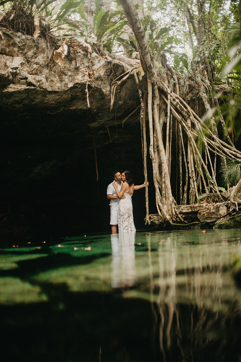 A couple stand together kissing in the water of a cenote in Mexico wearing a beautiful white dress and white shorts with a shirt for a Cenote Azul engagement photography session