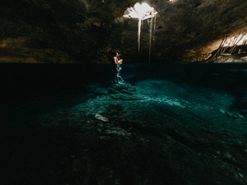 A couple stand together in the water of a cenote in Mexico wearing a white swimsuit and orange shorts for a Cenote Azul engagement photography session