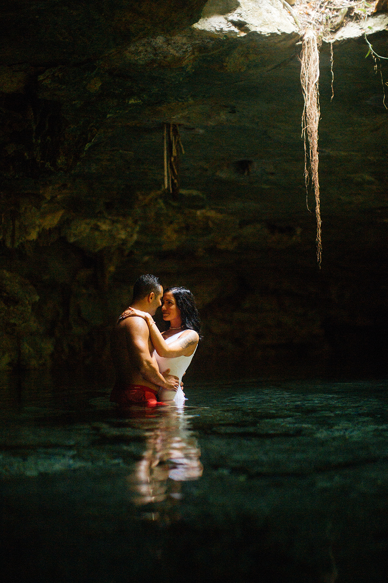 A couple stand together in the water of a cenote in Mexico wearing a white swimsuit and orange shorts for a Cenote Azul engagement photography session