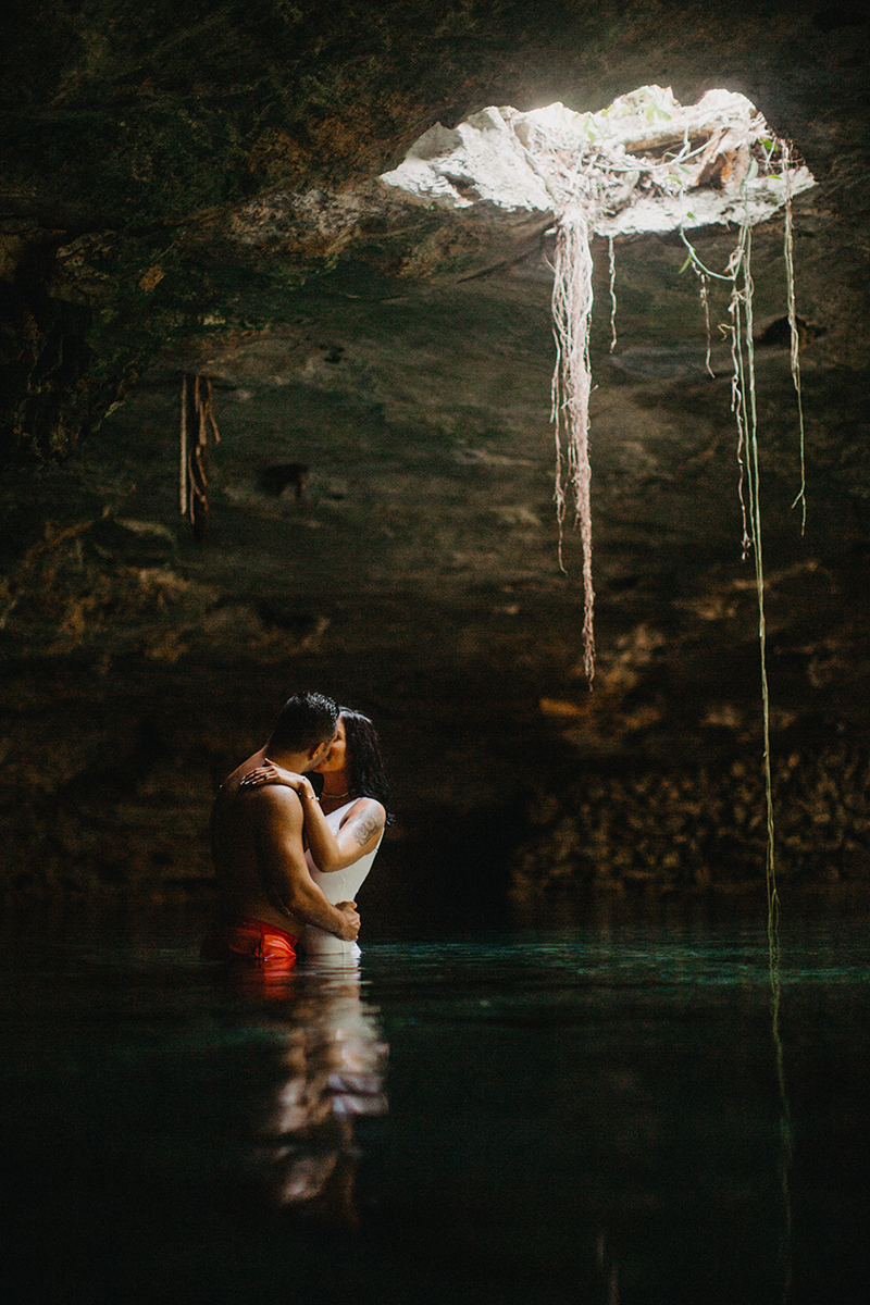 A couple stand together kissing in the water of a cenote in Mexico wearing a white swimsuit and orange shorts for a Cenote Azul engagement photography session