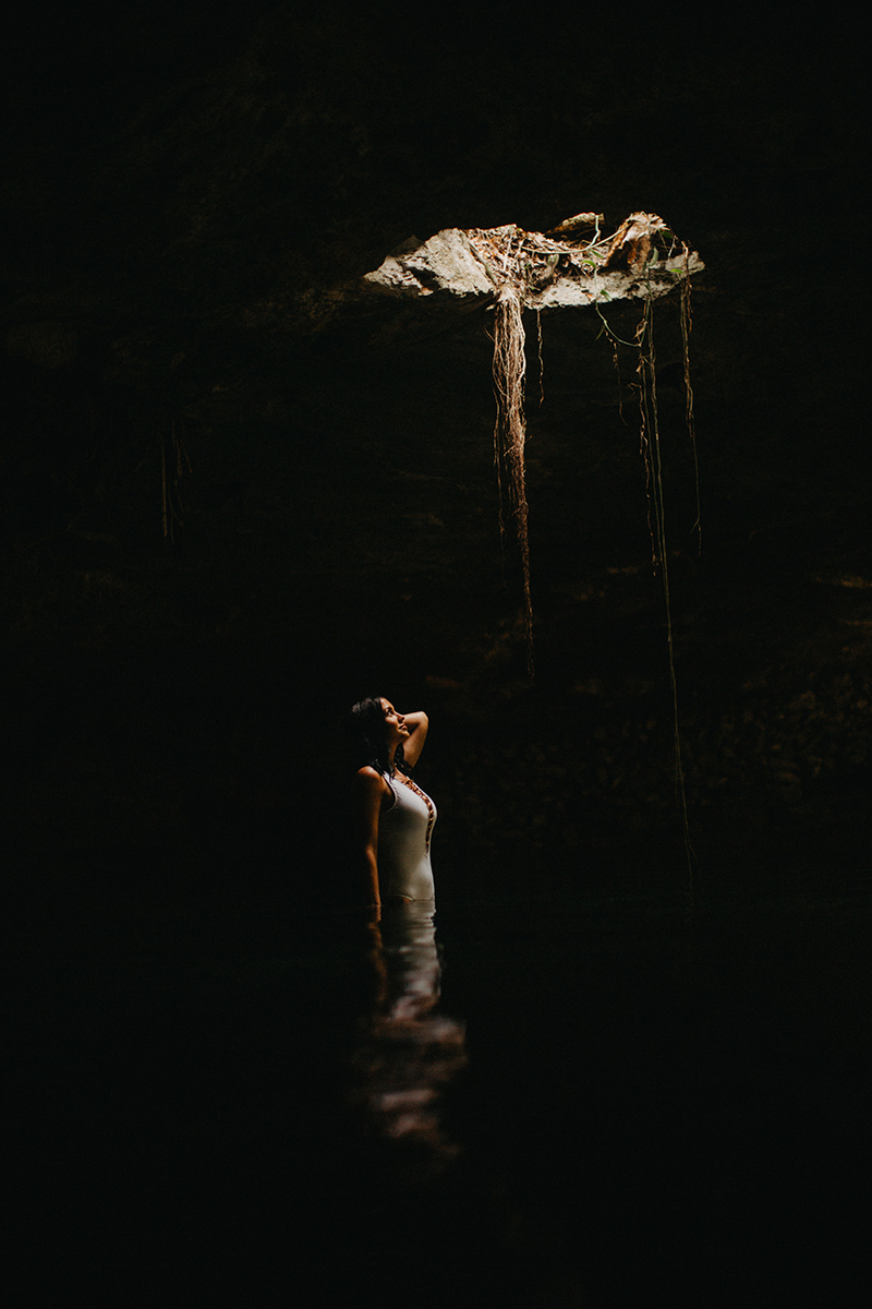 A beautiful woman poses in the water of a cenote in Mexico wearing a white swimsuit for a Cenote Azul engagement photography session