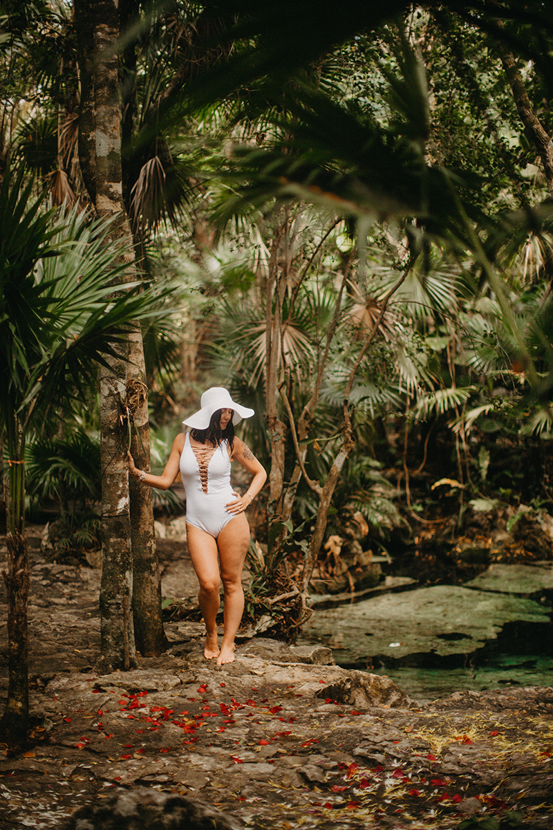 A beautiful woman poses near a cenote in Mexico wearing a white swimsuit and a beach hat for a Cenote Azul engagement photography sessionA beautiful woman poses near a cenote in Mexico wearing a white swimsuit and a beach hat for a Cenote Azul engagement photography session