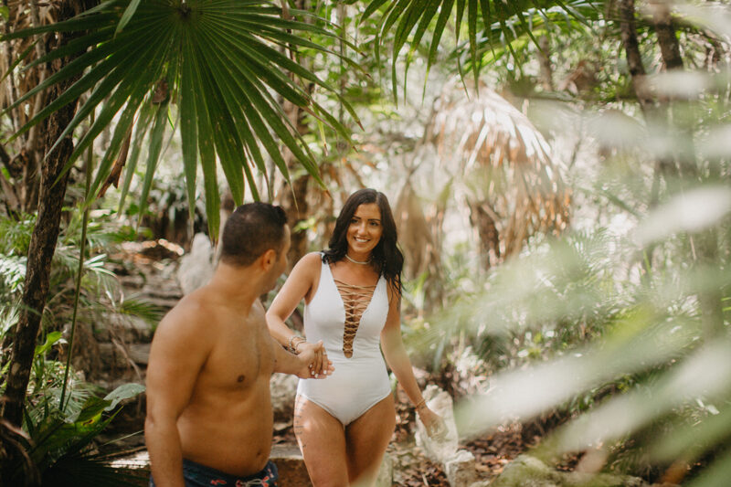 A couple walk together through the jungle near a cenote in Mexico wearing a white swimsuit and orange shorts for a Cenote Azul engagement photography session