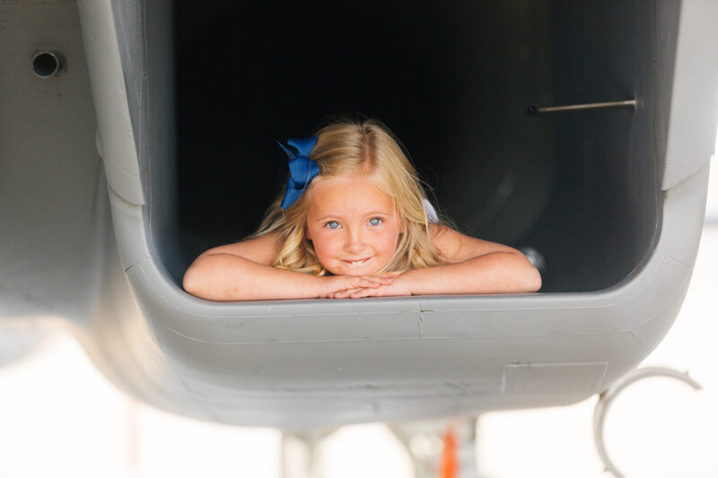 A girl laying down in the intake of an F-15 at Barnes Air National Guard Base wearing a dress for these F-15 fighter pilot family photos