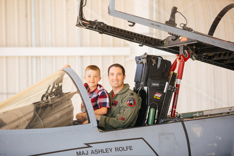 A pilot sits in the cockpit of an F-15 with his son on his lap at Barnes Air National Guard Base wearing a flight suit and a coordinated outfit for these F-15 fighter pilot family photos