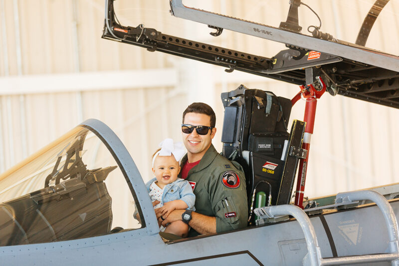 A pilot sits in the cockpit of an F-15 with his daughter on his lap at Barnes Air National Guard Base wearing a flight suit and a coordinated outfit for these F-15 fighter pilot family photos
