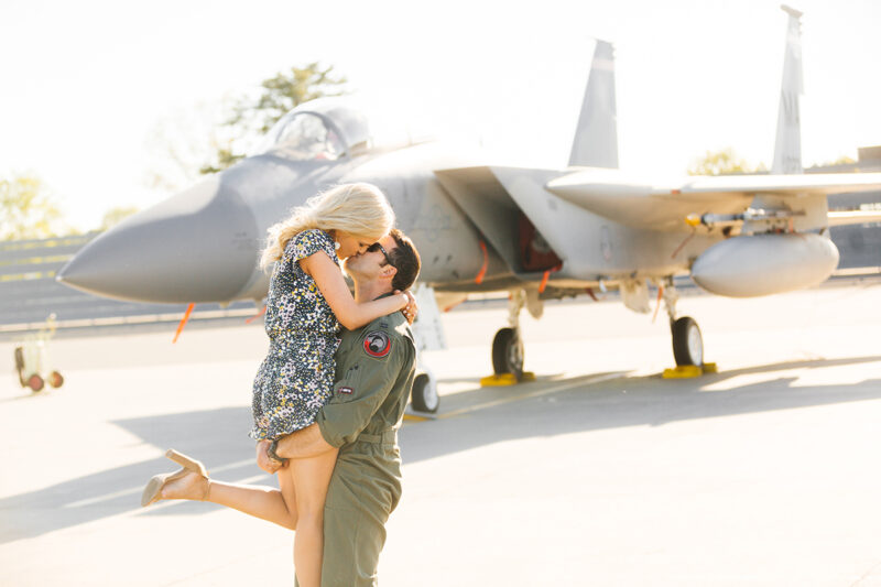 A pilot lifts his wife up and kisses her in front of an F-15 at Barnes Air National Guard Base wearing a flight suit and a coordinated outfit for these F-15 fighter pilot family photos