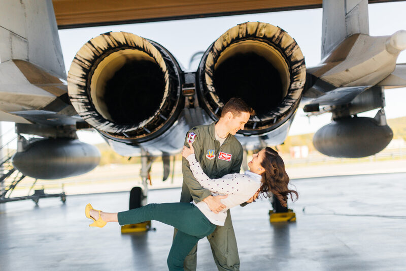 A pilot and his wife stand together as he dips her behind an F-15 at Barnes Air National Guard Base wearing a flight suit and a coordinated outfit for these F-15 fighter pilot family photos