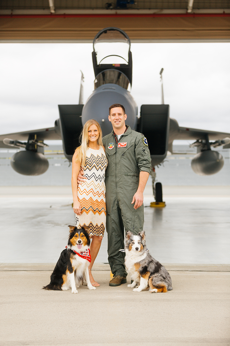 A pilot and his wife stand together with their dogs in front an F-15 at Barnes Air National Guard Base wearing a flight suit and coordinated outfits for these F-15 fighter pilot family photos