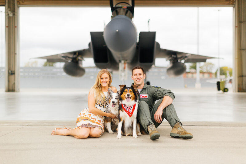 A pilot and his wife sit together with their dogs in front an F-15 at Barnes Air National Guard Base wearing a flight suit and coordinated outfits for these F-15 fighter pilot family photos