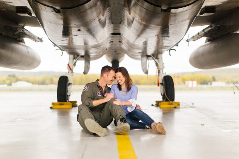 A pilot and his wife sit together under an F-15 at Barnes Air National Guard Base wearing a flight suit and a coordinated outfit for these F-15 fighter pilot family photos