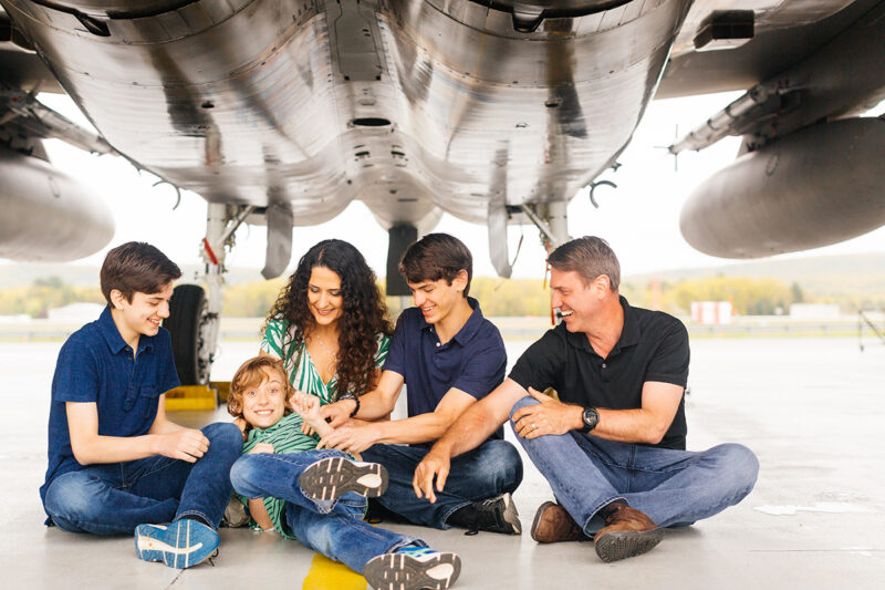 A family sits under an F-15 holding each other close tickling each other at Barnes Air National Guard Base wearing coordinated outfits for these F-15 fighter pilot family photos