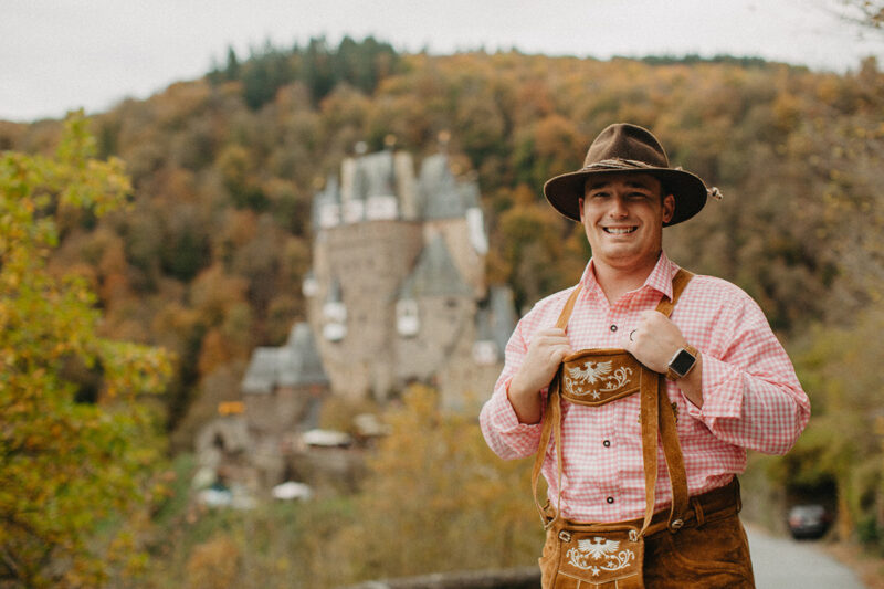 A handsome young man poses on the road to Burg Eltz wearing traditional lederhosen for these Eltz Castle couples photos in Germany