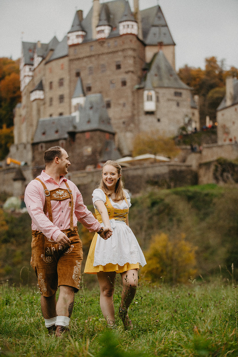 A couple walk together holding hands in a field near Burg Eltz wearing a traditional dirndl and lederhosen for these Eltz Castle couples photos in Germany