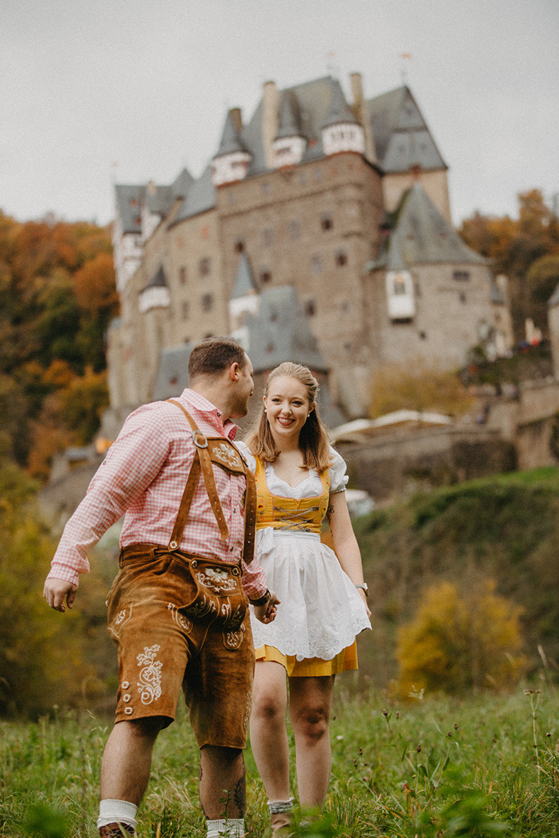A couple walk together holding hands in a field near Burg Eltz wearing a traditional dirndl and lederhosen for these Eltz Castle couples photos in Germany