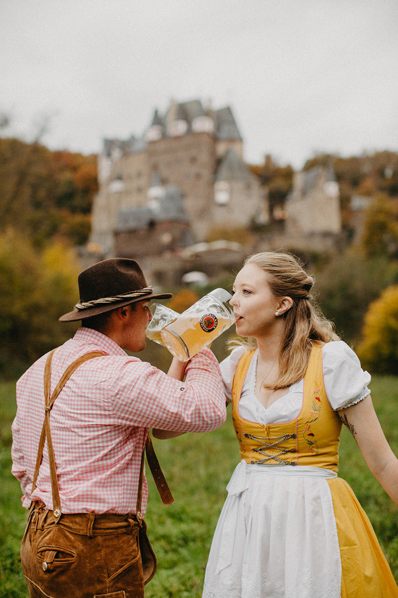 A couple drink together with mugs in a field near Burg Eltz wearing a traditional dirndl and lederhosen for these Eltz Castle couples photos in Germany