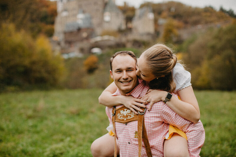 A couple embrace as he gives her a piggyback ride in a field near Burg Eltz wearing a traditional dirndl and lederhosen for these Eltz Castle couples photos in Germany