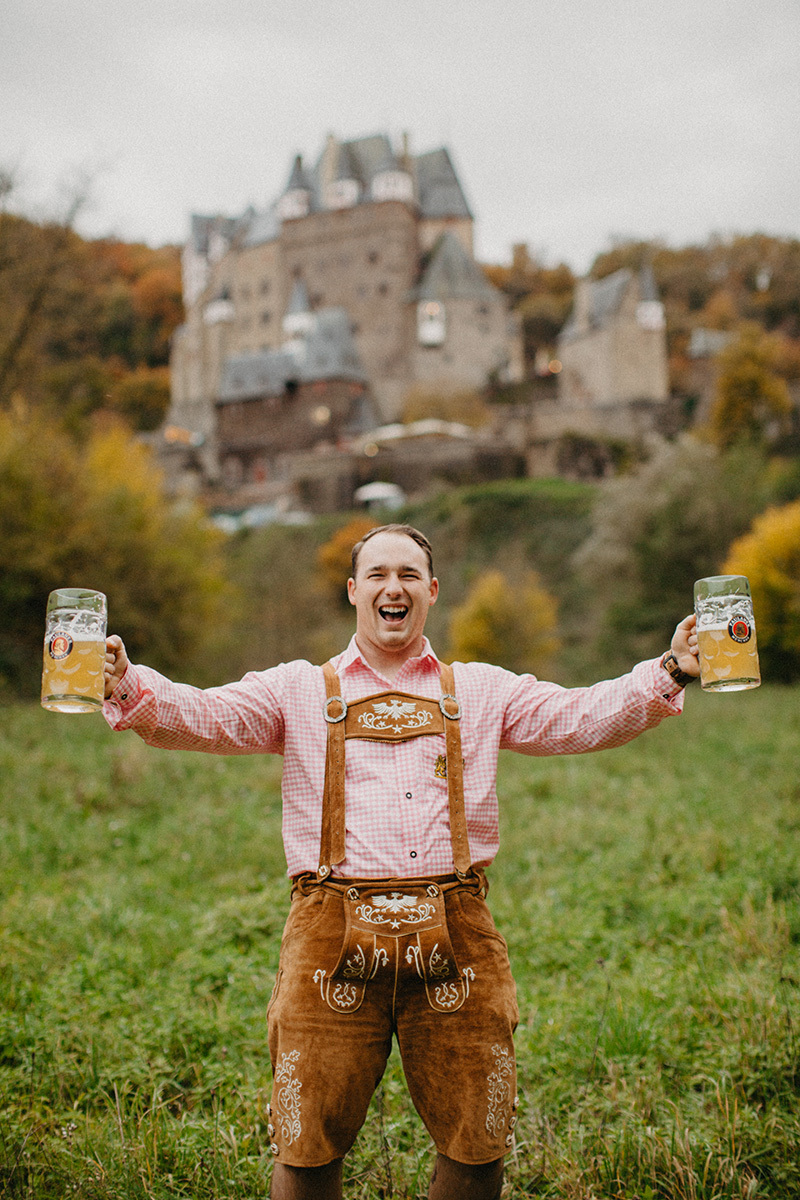 A handsome young man holds beer in a field near Burg Eltz wearing traditional lederhosen for these Eltz Castle couples photos in Germany