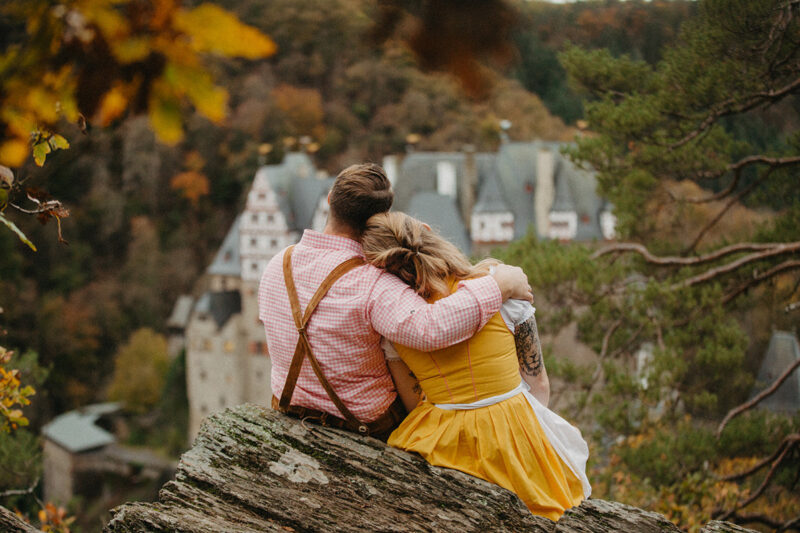 A couple embrace sitting on a ridge near Burg Eltz wearing a traditional dirndl and lederhosen for these Eltz Castle couples photos in Germany