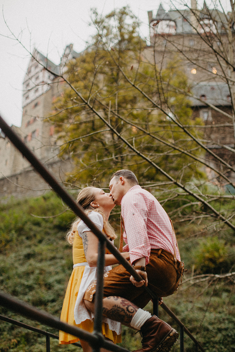 A couple kiss on a bridge near Burg Eltz wearing a traditional dirndl and lederhosen for these Eltz Castle couples photos in Germany