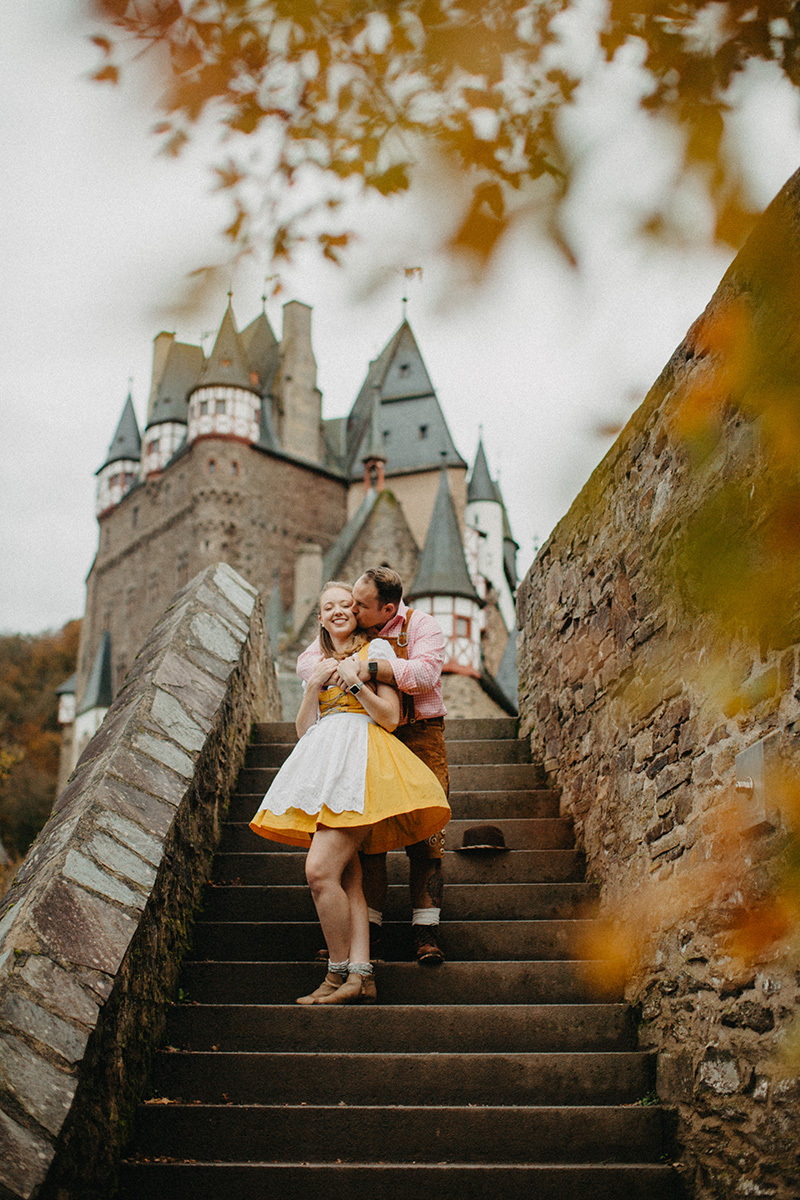 A couple embraces on stairs near Burg Eltz wearing a traditional dirndl and lederhosen for these Eltz Castle couples photos in Germany