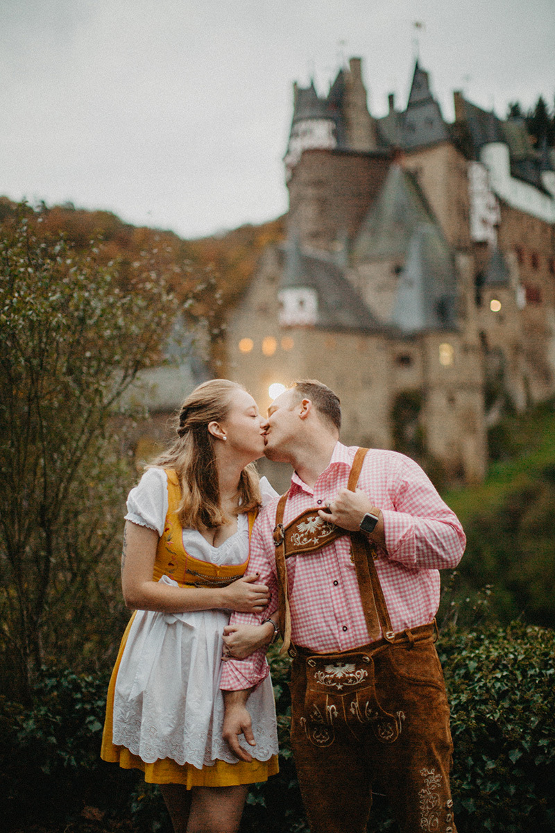 A couple holds hands and kiss in front of Burg Eltz wearing a traditional dirndl and lederhosen for these Eltz Castle couples photos in Germany
