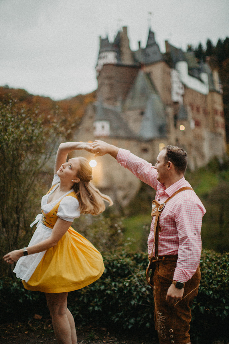 A couple dances in front of Burg Eltz wearing a traditional dirndl and lederhosen for these Eltz Castle couples photos in Germany