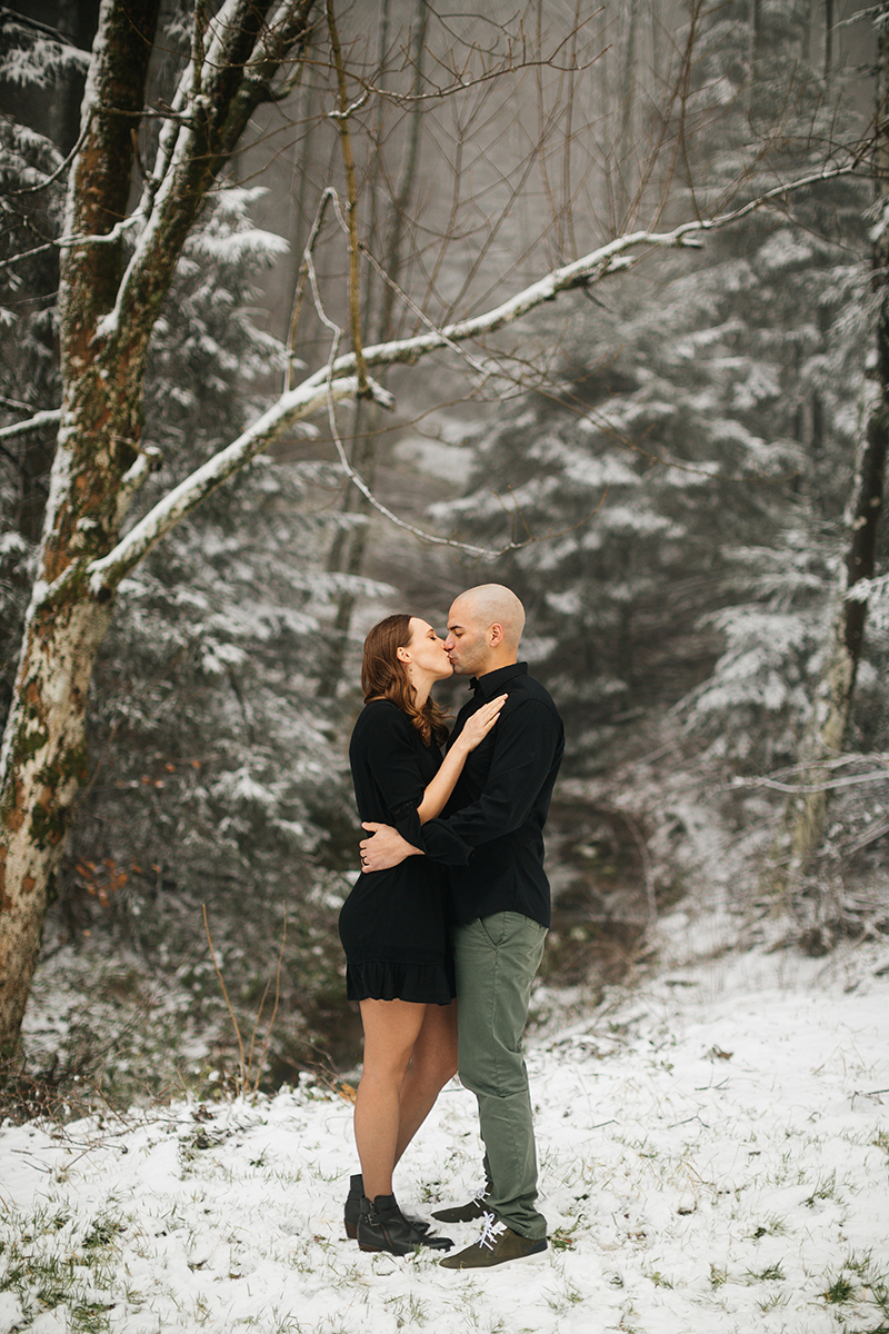A couple kiss while they embrace in a snow covered forest in Germany wearing a beautiful black dress and black button up shirt for a Black Forest engagement photography session