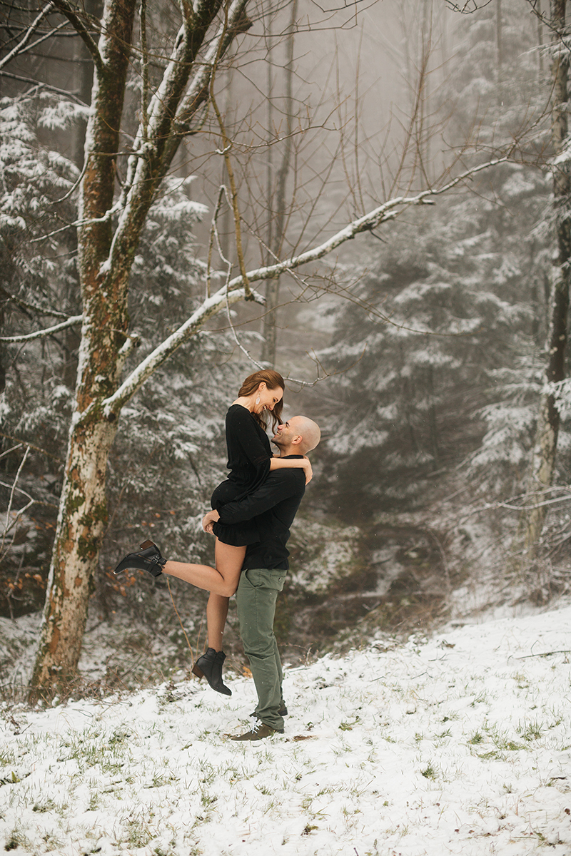 A couple embrace as he lifts her up in a snow covered forest in Germany wearing a beautiful black dress and black button up shirt for a Black Forest engagement photography session