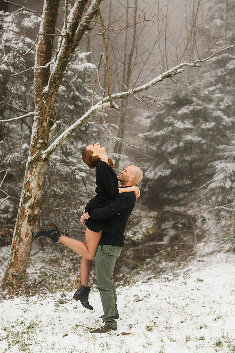 A couple embrace as he lifts her up in a snow covered forest in Germany wearing a beautiful black dress and black button up shirt for a Black Forest engagement photography session