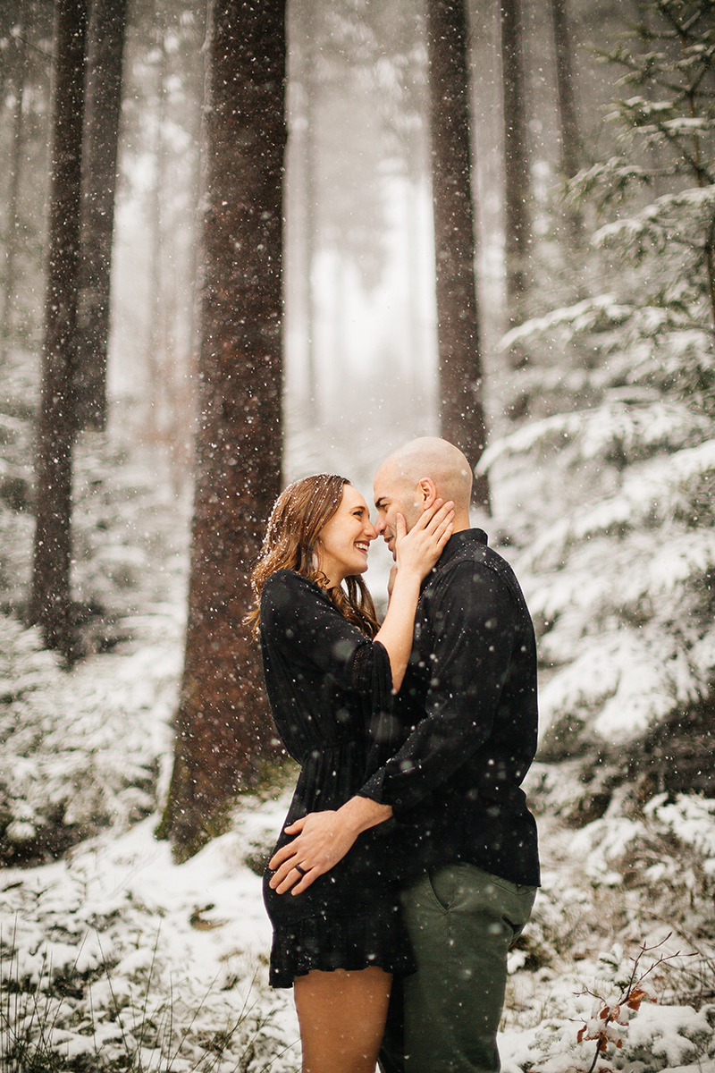 A couple embrace in a snow covered forest in Germany wearing a beautiful black dress and black button up shirt for a Black Forest engagement photography session